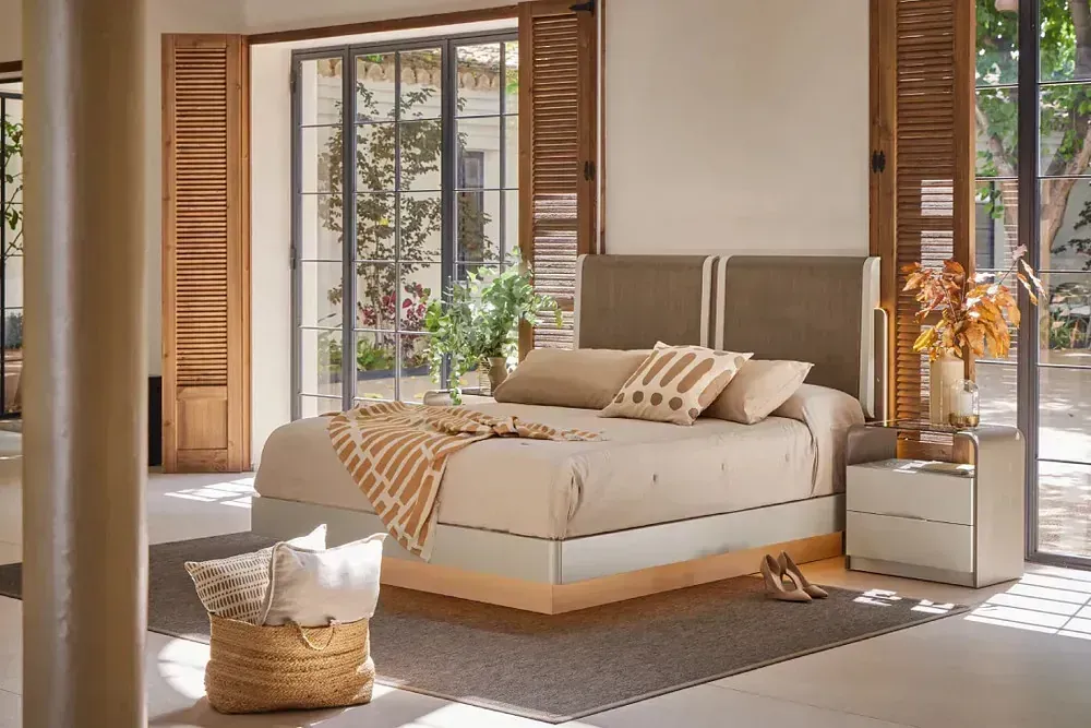 Dormitorio con cama, mesita de noche, persianas abiertas y cojines decorativos en tonos neutros.