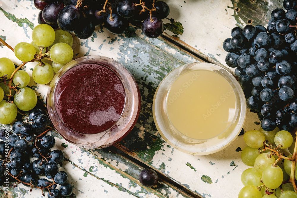 Zwei Gläser Saft, eines lila, eines gelb, umgeben von grünen und lila Trauben auf einer Holzoberfläche.