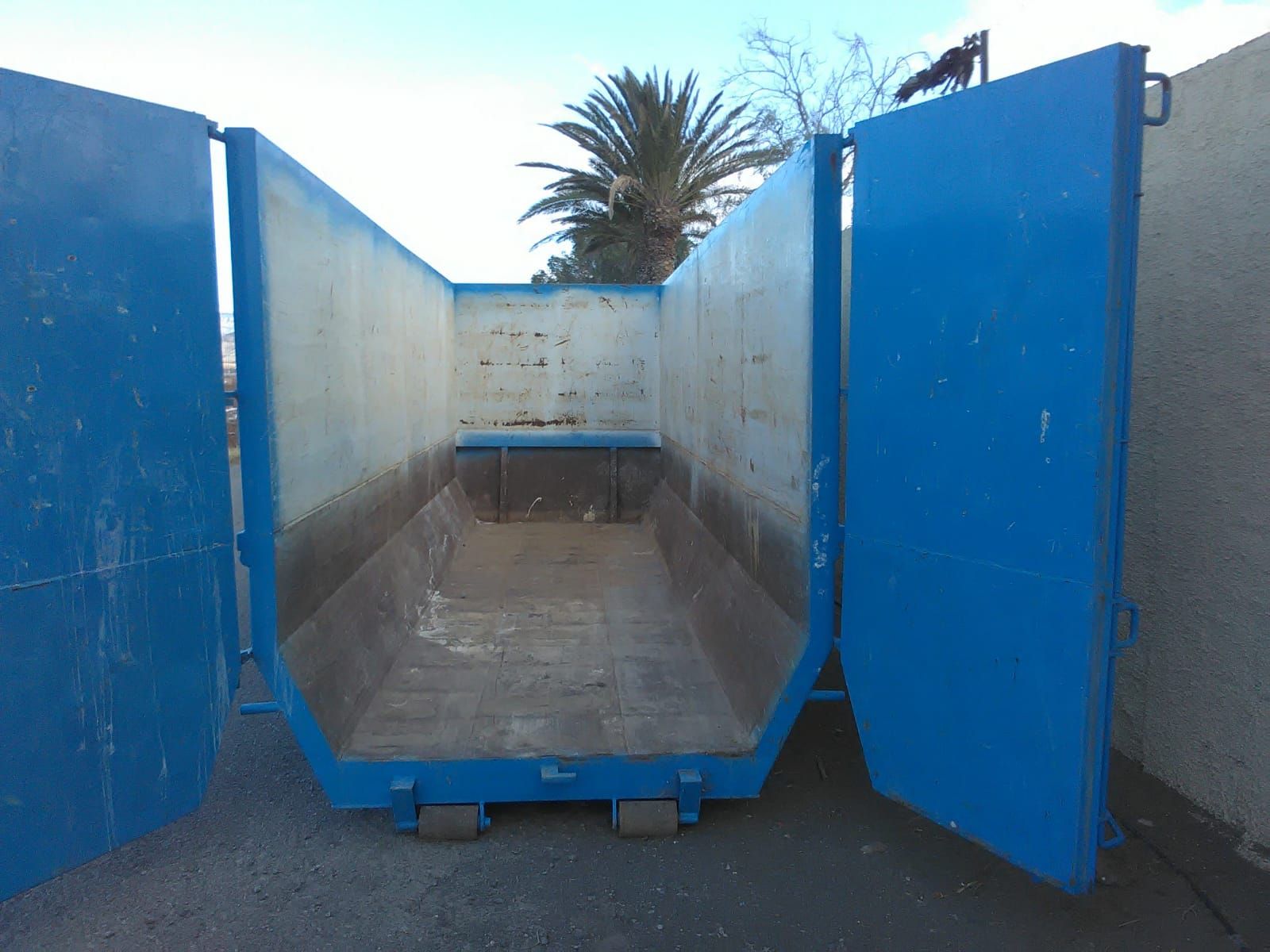 Un contenedor de basura azul con las puertas abiertas y una palmera al fondo.