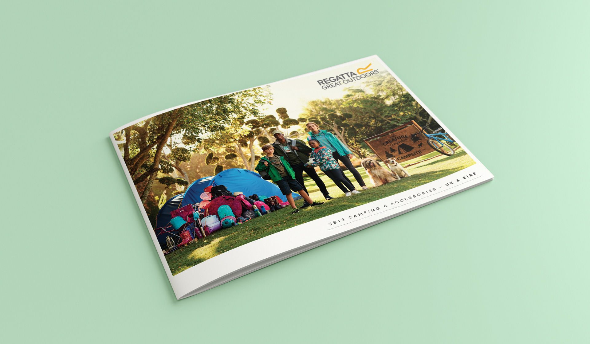 A brochure mockup lying on a light green surface displays a photo of a group camping together in a grassy, wooded area.