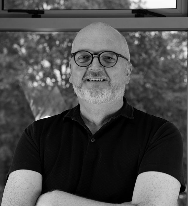 A smiling person with glasses and a beard, wearing a polo shirt, standing in front of a window with a blurred leafy background.