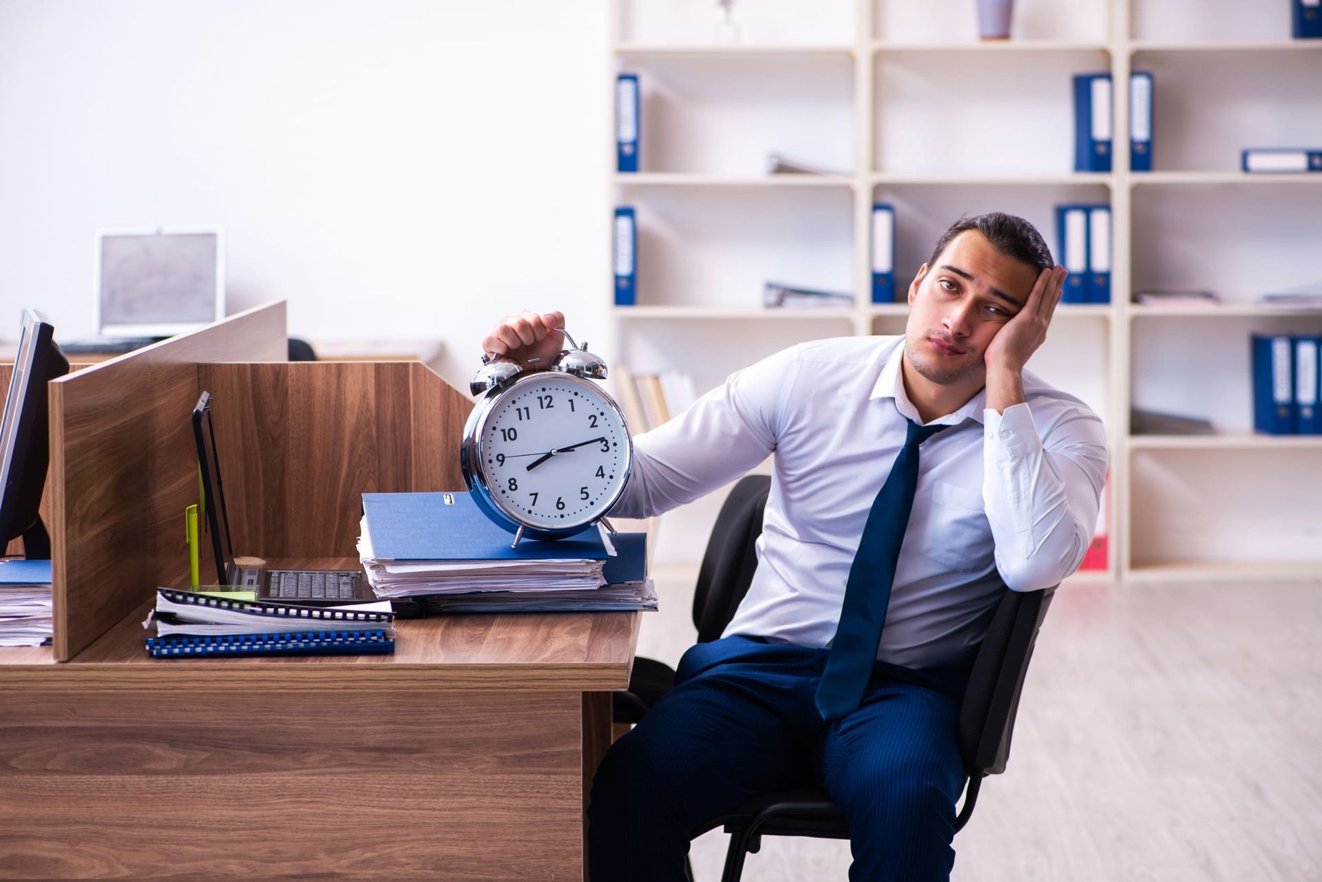 Un hombre está sentado en un escritorio en una oficina sosteniendo un reloj despertador.