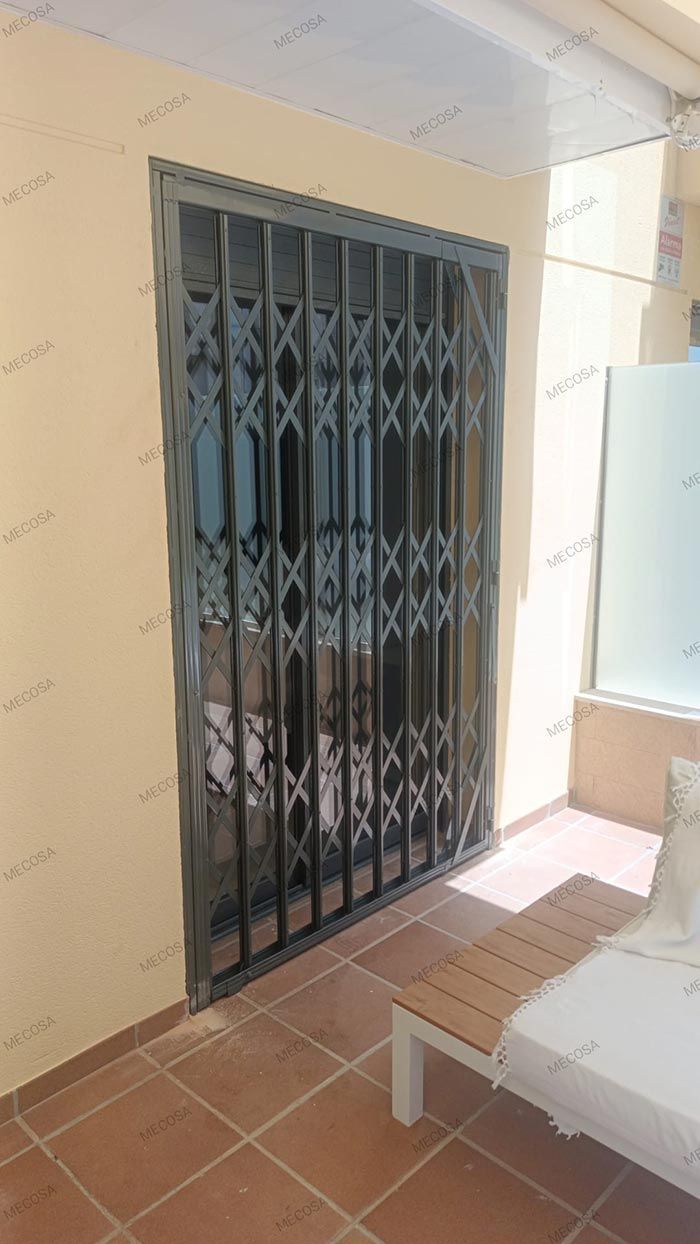 Una verja de seguridad de metal negro se encuentra en un porche con baldosas, junto a un edificio blanco.