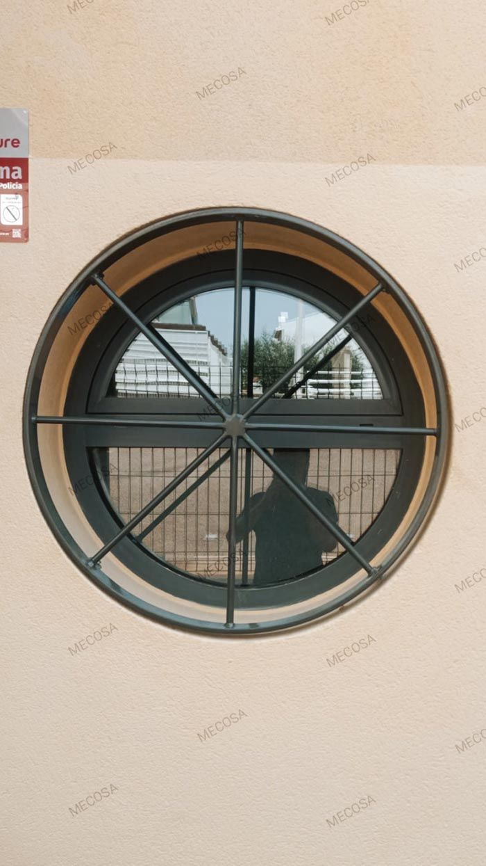 Ventana redonda con travesaños negros en una pared beige, que deja ver un interior borroso con una silla y reflejos en el exterior.
