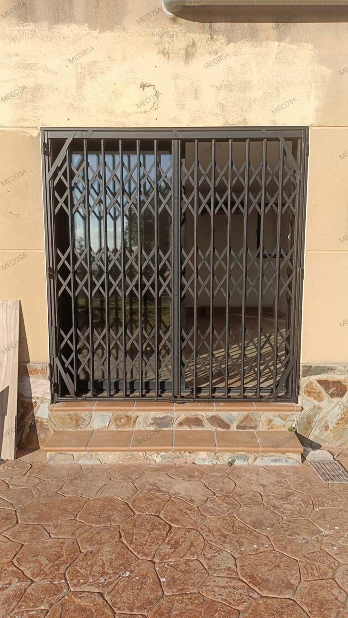 Puerta de seguridad corrediza sobre una ventana. La puerta es negra y la ventana refleja árboles. Está ubicada en un edificio de color claro.