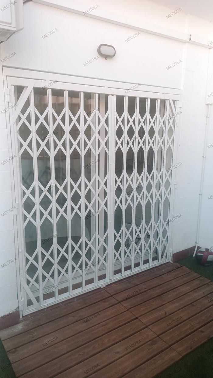Una verja de seguridad de metal blanco cubre la entrada de una terraza de madera junto a una pared blanca.