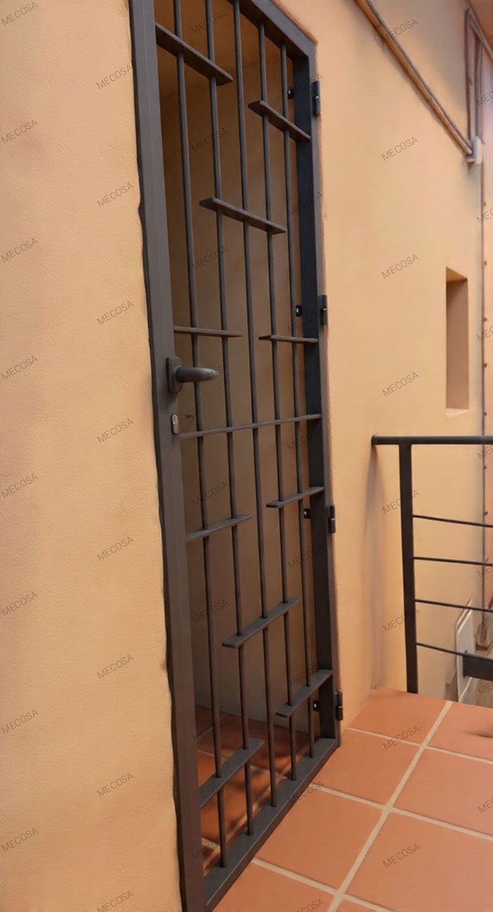 Puerta de seguridad metálica en una pared exterior de color melocotón, junto a un balcón con azulejos.