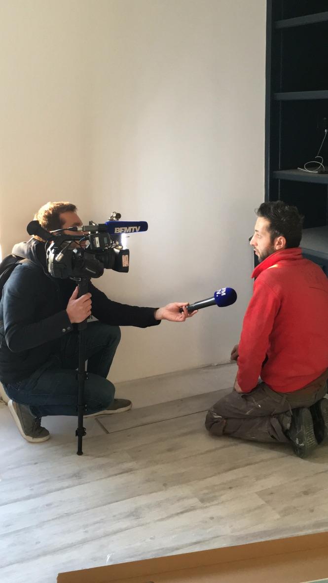 Un caméraman interviewe une personne en chemise rouge, agenouillée au sol. L'interview a lieu en intérieur.