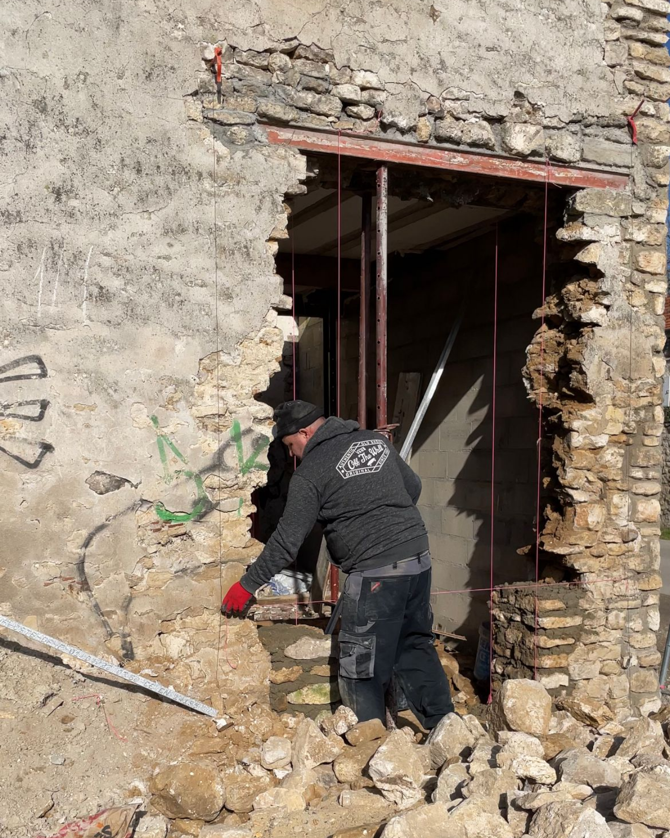 Un homme travaille sur l'encadrement d'une porte d'immeuble endommagée. Mur de pierre effondré, supports métalliques, débris au sol.