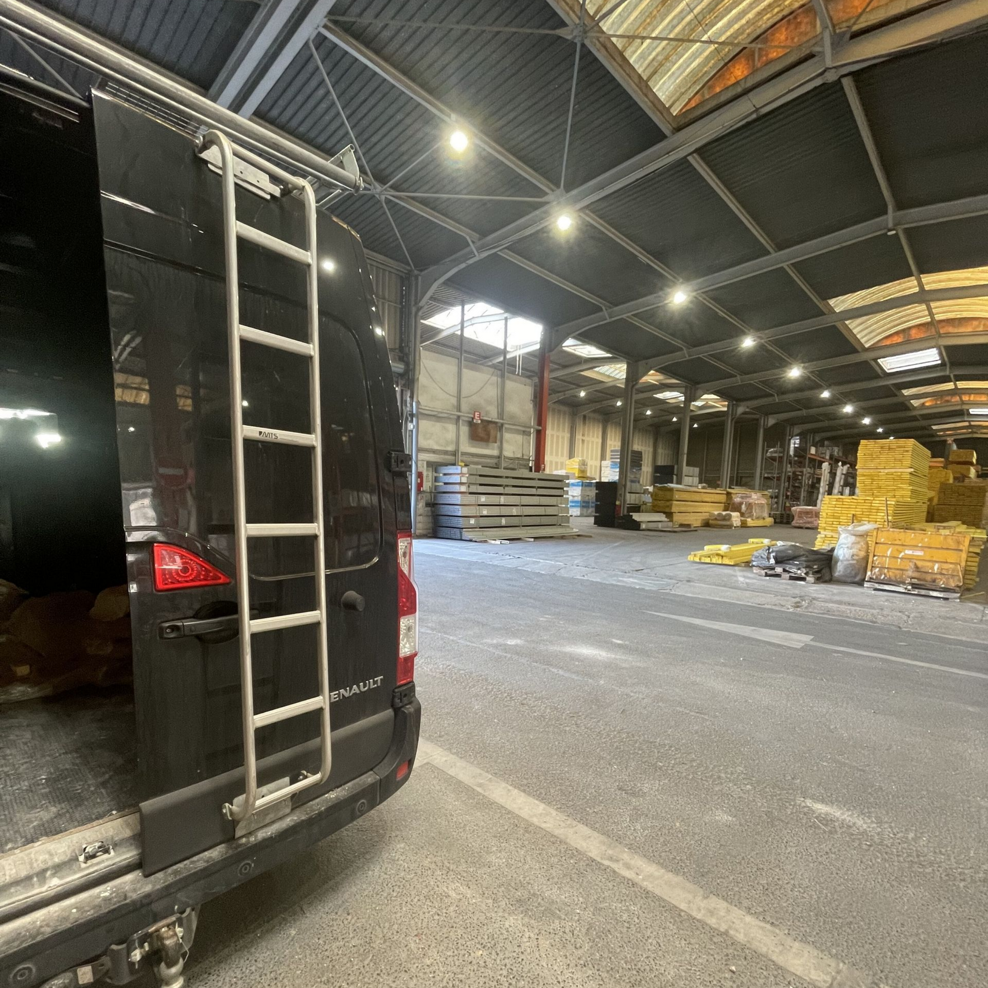Fourgonnette noire, porte ouverte, stationnée à l'intérieur d'un grand entrepôt. Échelle fixée à l'arrière.