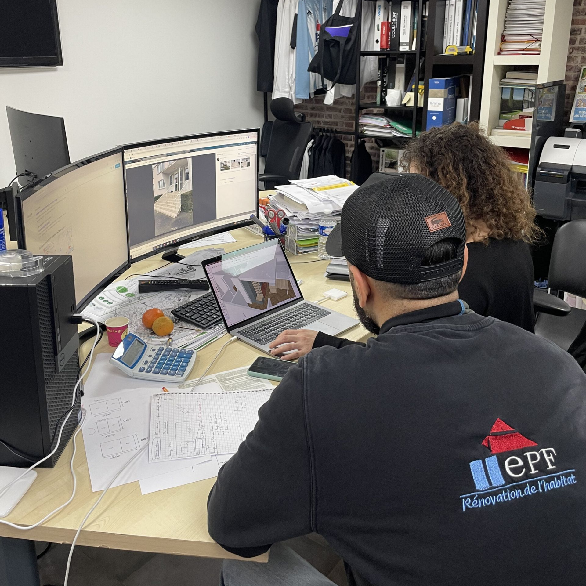 Deux personnes travaillent à un bureau équipé d'ordinateurs. Aménagement de bureau avec des documents, une imprimante et un mur de briques.