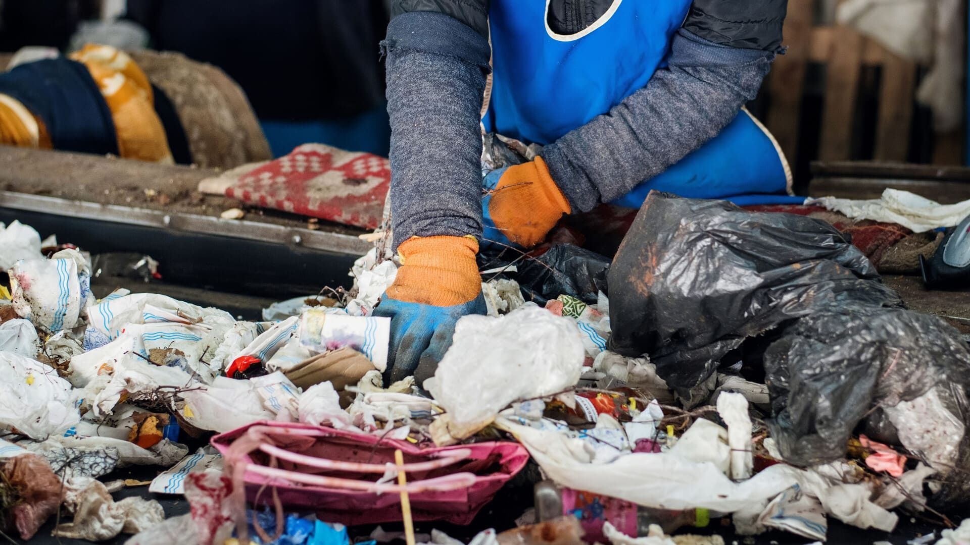 Una persona con delantal azul y guantes naranjas clasifica una pila de residuos plásticos
