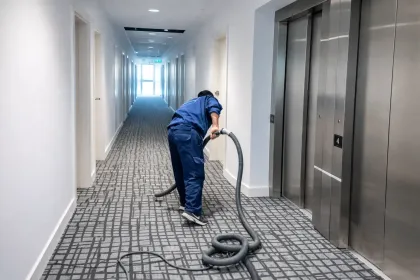 Una persona vestida con ropa de trabajo azul aspira la alfombra de un pasillo junto a un conjunto de puertas