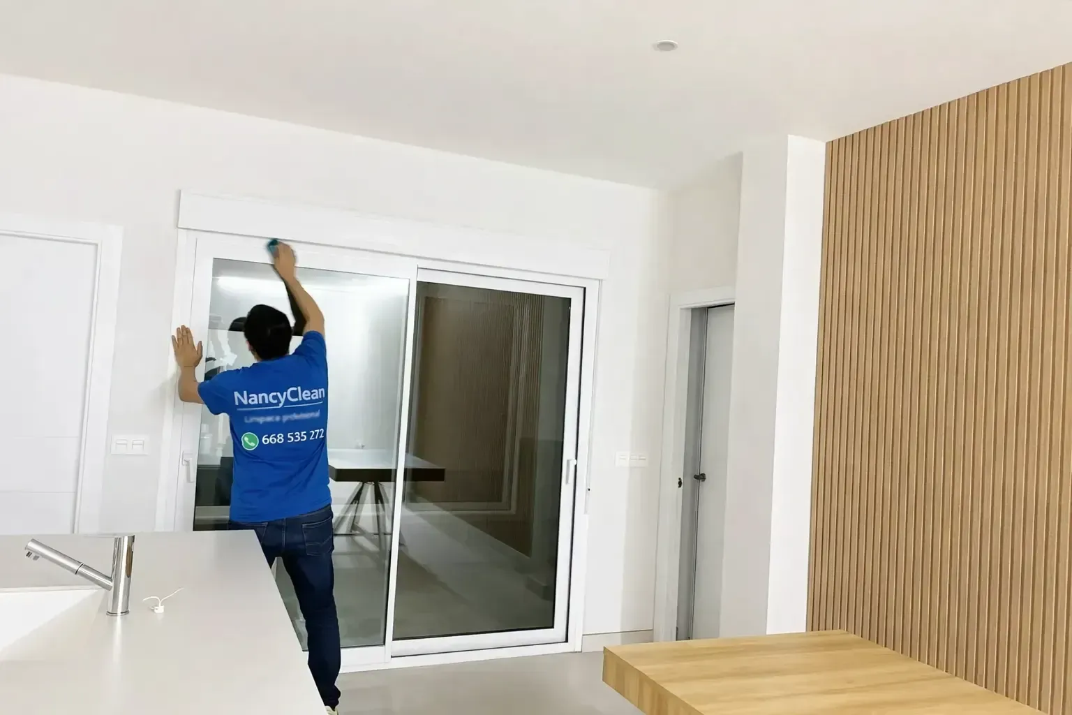 Una persona con una camisa azul limpia el marco superior de una puerta corrediza de vidrio blanca