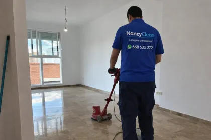 Una persona friega el suelo de la entrada de un edificio con puertas de cristal enmarcadas en madera clara.