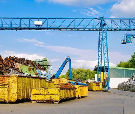 Un patio de reciclaje de chatarra con contenedores amarillos, una grúa azul y pilas de metal contra un cielo azul.