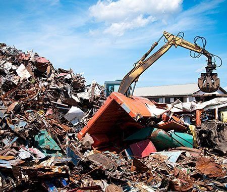 Excavadora levantando chatarra en un patio de reciclaje bajo un cielo azul.
