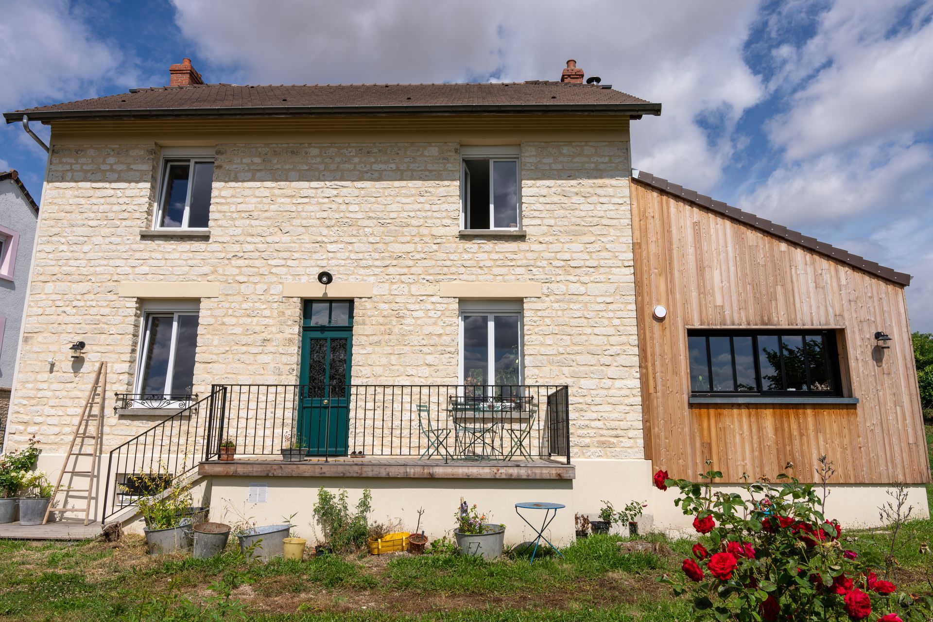 Maison en pierre avec une porte turquoise et un balcon, à côté d’une extension moderne à bardage en bois.