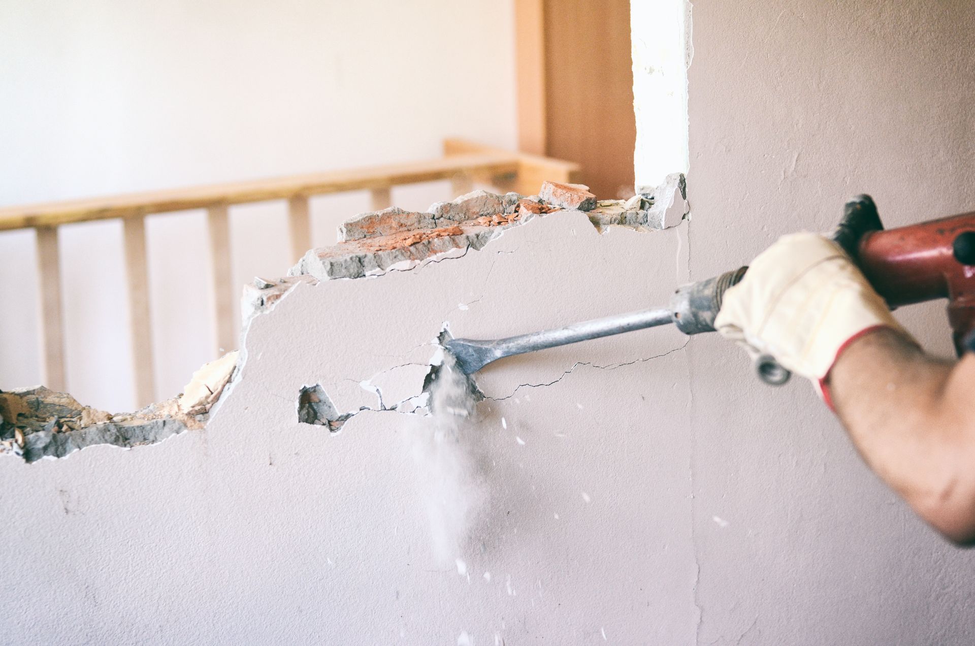 Une main gantée décolle le plâtre d’un mur endommagé à l’aide d’un outil, révélant l’intérieur sous-jacent.