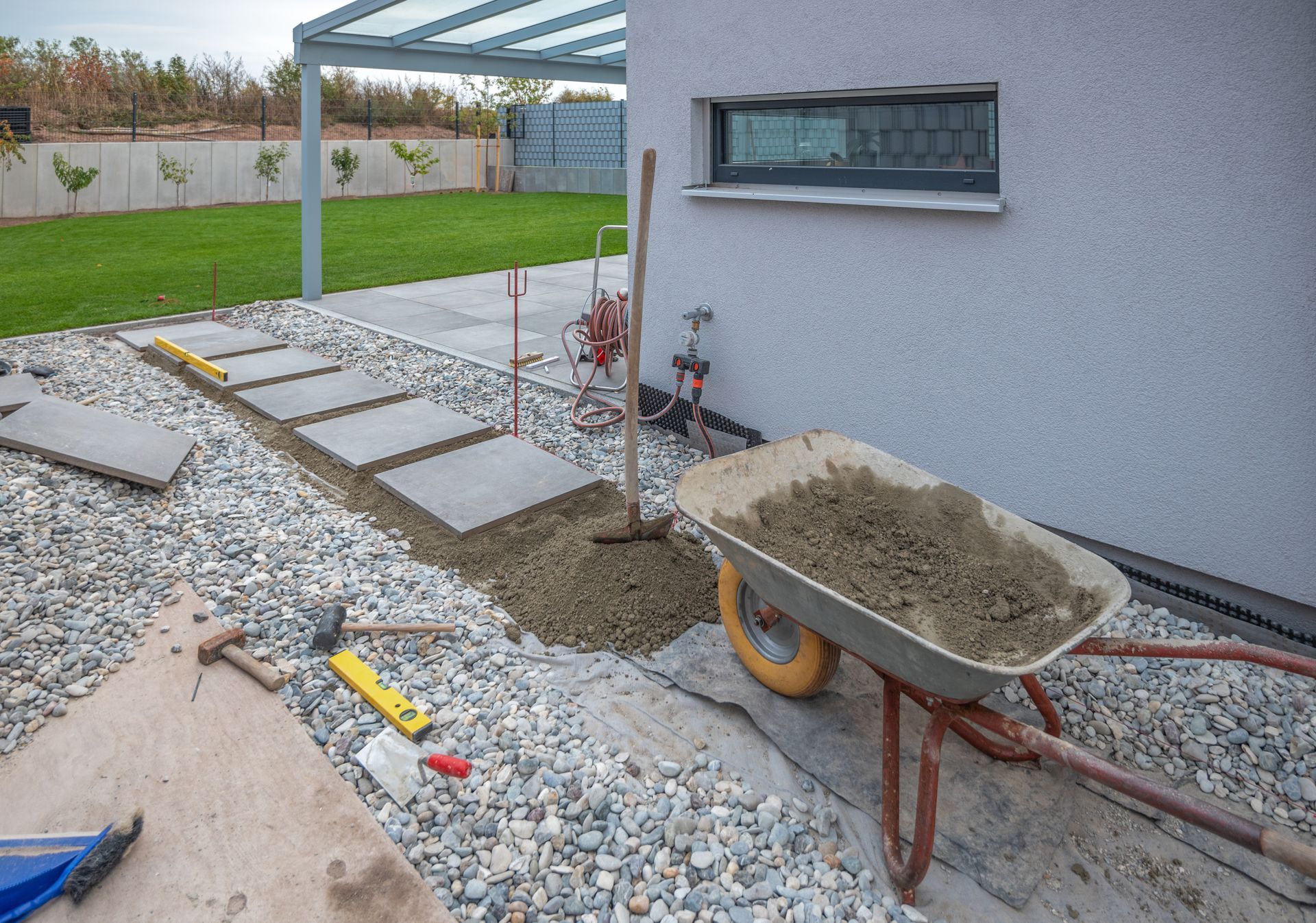 Une brouette remplie de gravier près du mur d’une maison, avec des dalles de pavage et une structure d’abri.