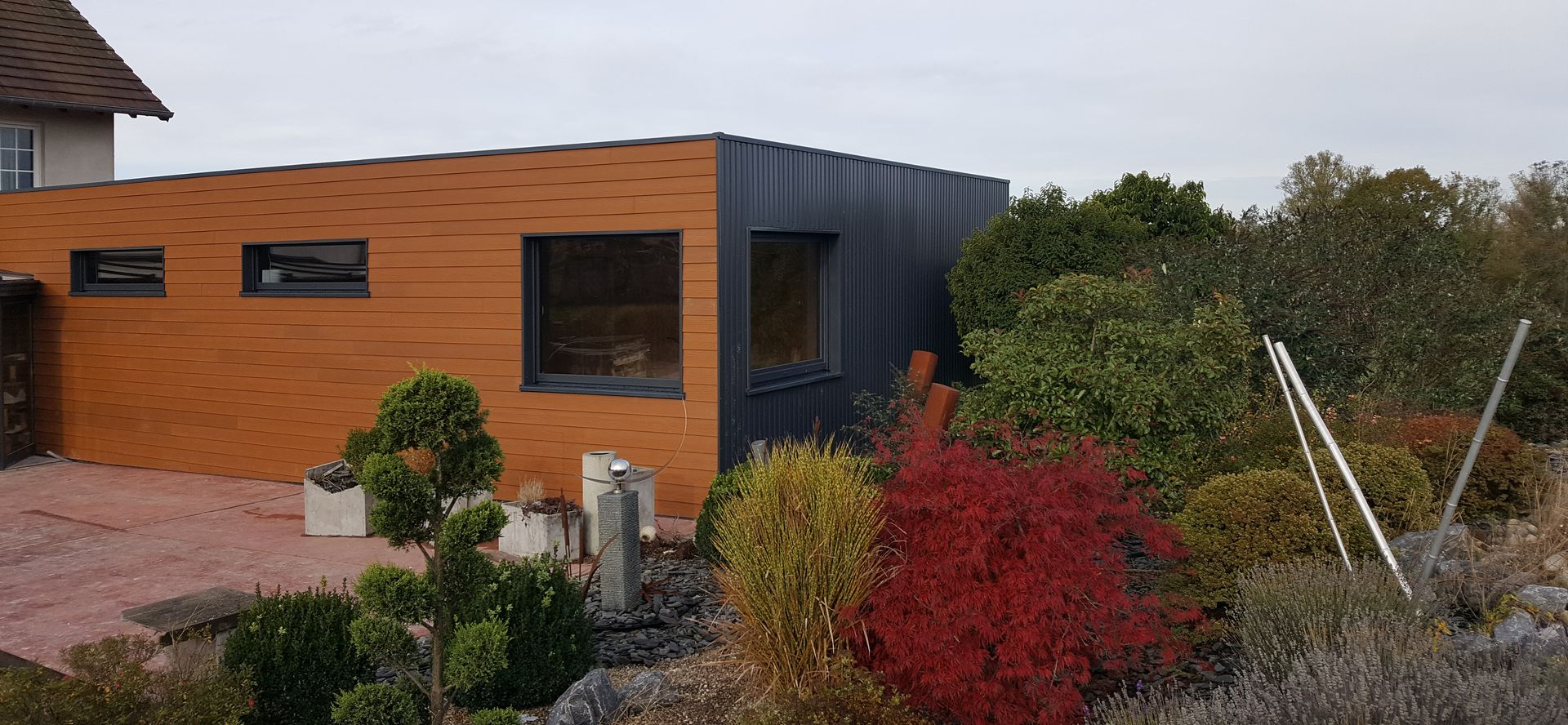 Un bâtiment moderne aux façades en bois brun et en métal foncé, entouré d'une végétation colorée et d'un patio en briques rouges.