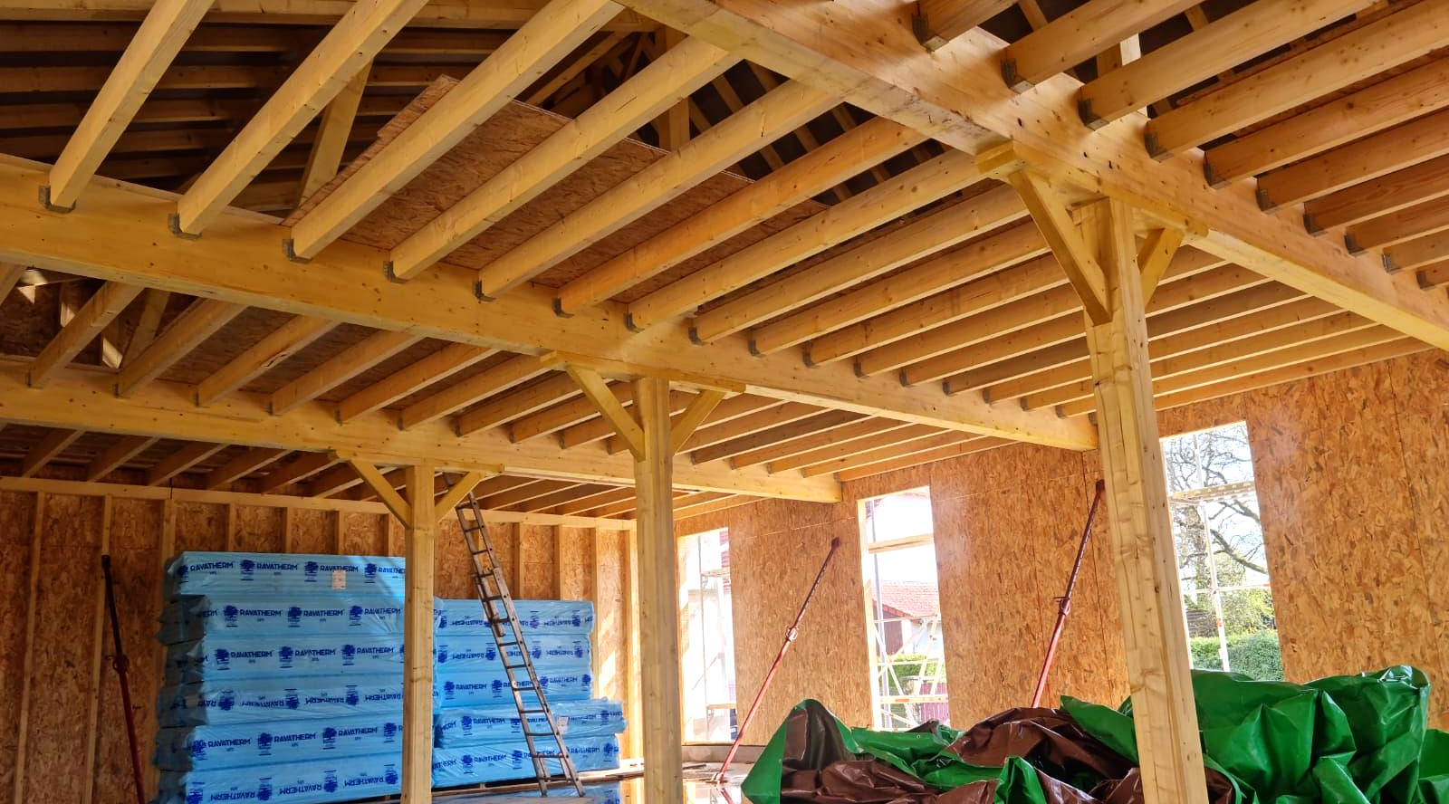 La charpente en bois d'un bâtiment en construction, sous un soleil éclatant et un ciel bleu visible.