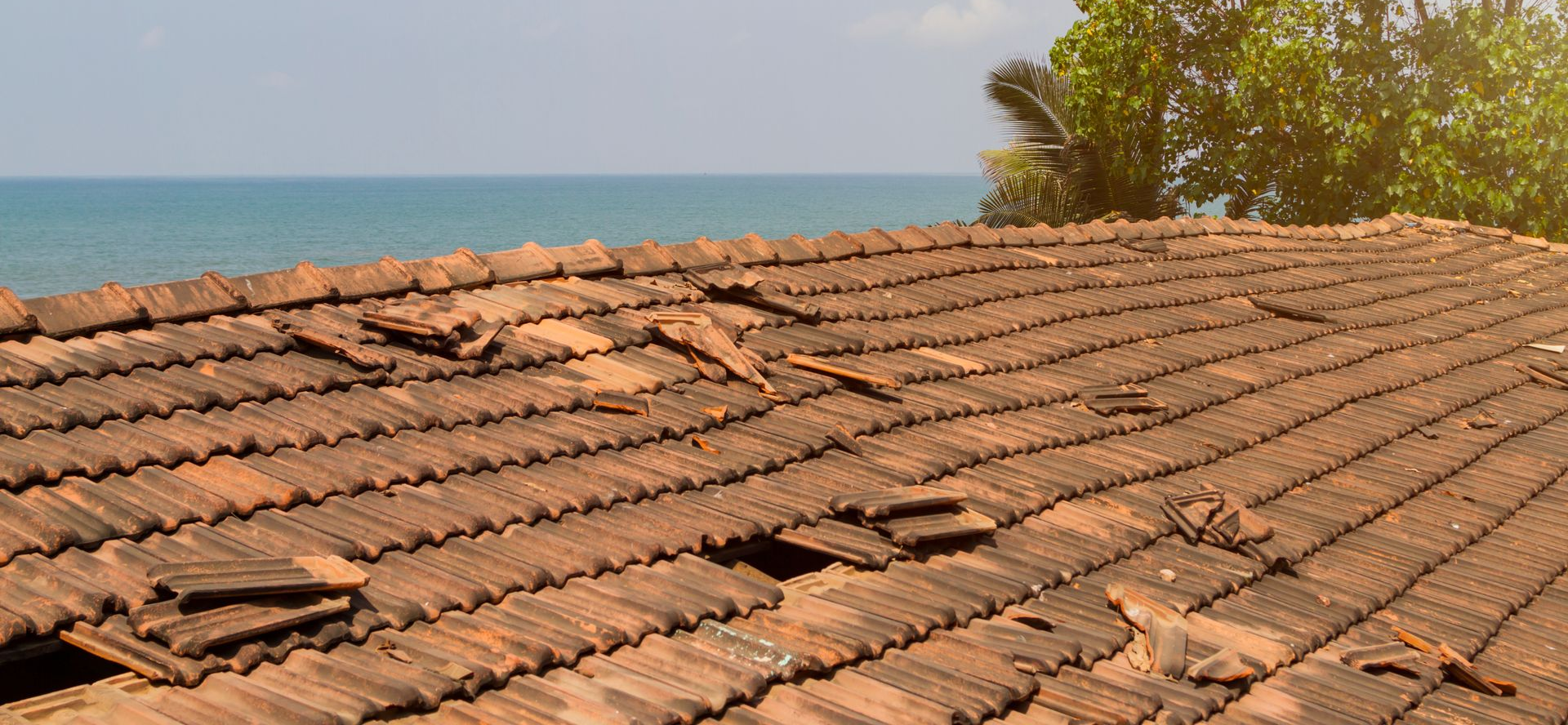 Un toit de tuiles en terre cuite délabré, aux tuiles manquantes, donnant sur l'océan et la verdure par une journée ensoleillée.