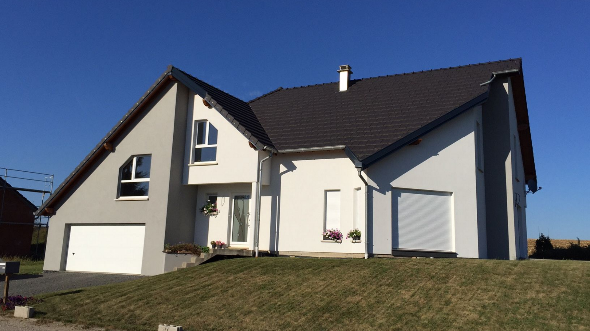 Maison moderne à deux étages avec façade grise et blanche, garage et toit foncé, par une journée ensoleillée.