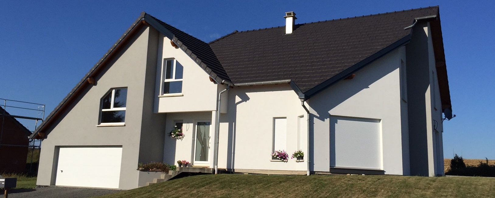 Une maison moderne à deux étages avec une porte de garage blanche et un revêtement extérieur gris, par une journée ensoleillée.