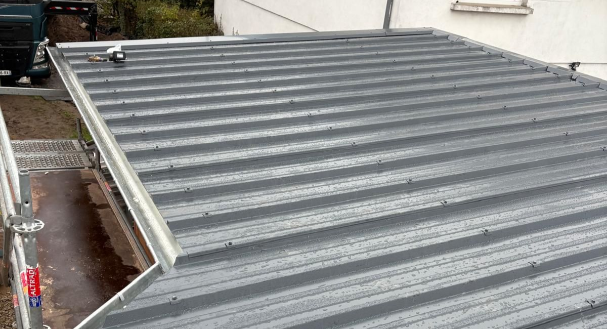 Toit en tôle ondulée grise avec une gouttière et un petit bâtiment en arrière-plan.