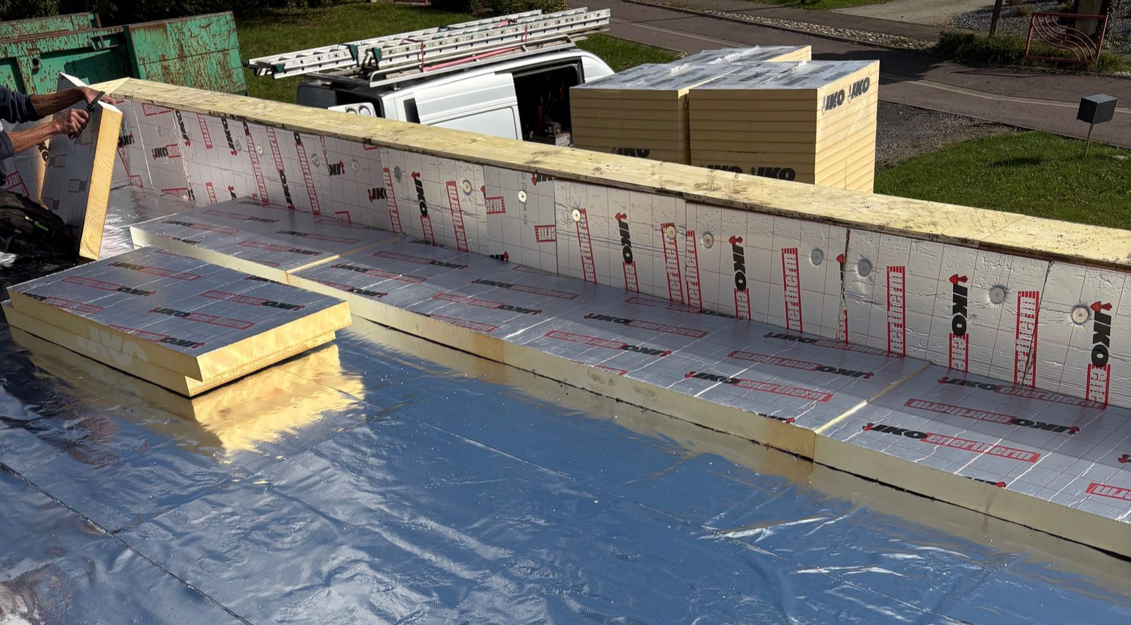 Chantier de construction avec pose d'isolant sur un toit. On aperçoit du papier aluminium, des panneaux isolants jaunes et blancs, ainsi qu'une charpente en bois.