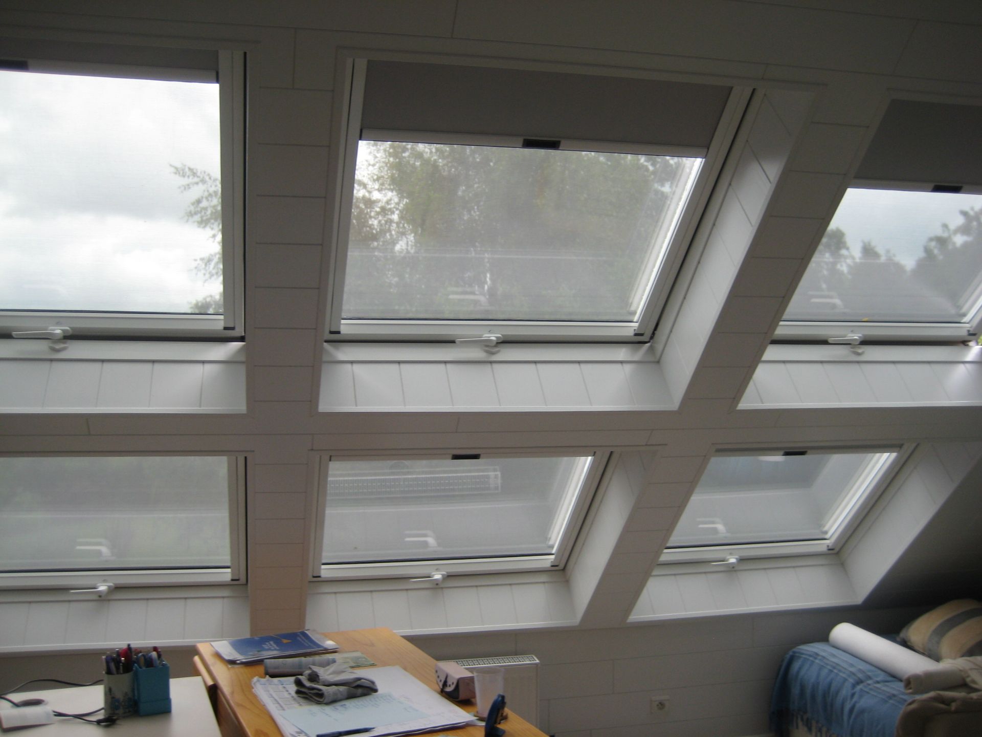 Six puits de lumière, percés dans un plafond à panneaux blancs, s'ouvrent sur un ciel nuageux. Un bureau se trouve en dessous.