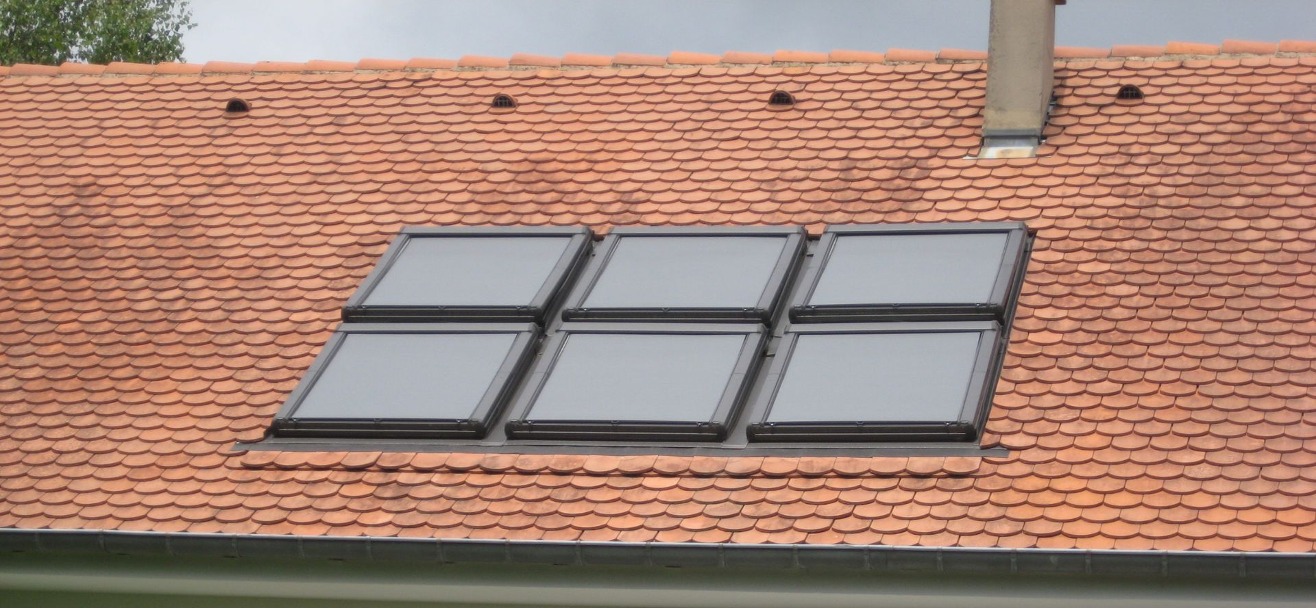 Un toit de tuiles en terre cuite percé de six puits de lumière et une cheminée, par temps nuageux.