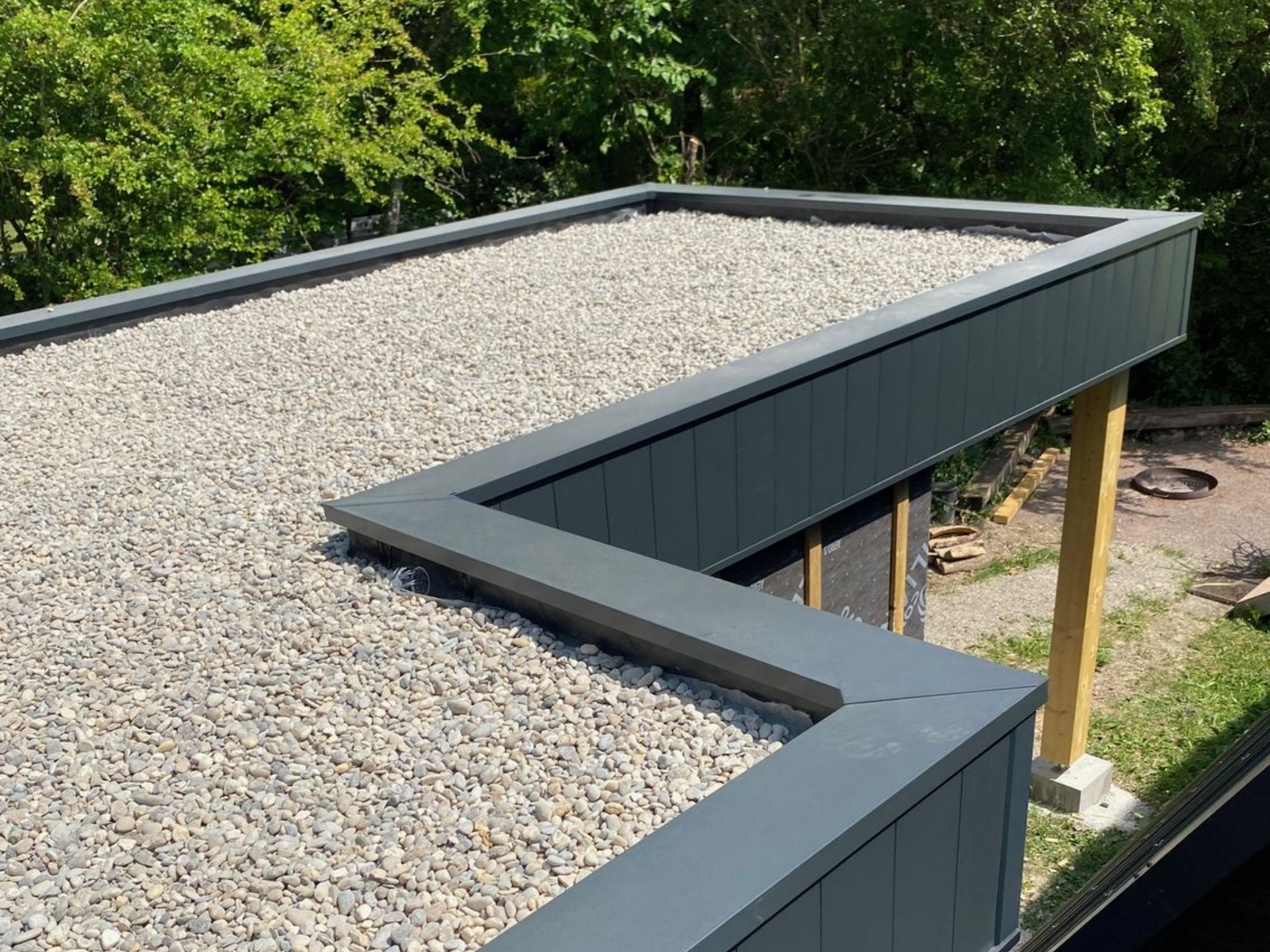 Toit plat recouvert de petits cailloux avec une bordure métallique grise. Arbres verts en arrière-plan.