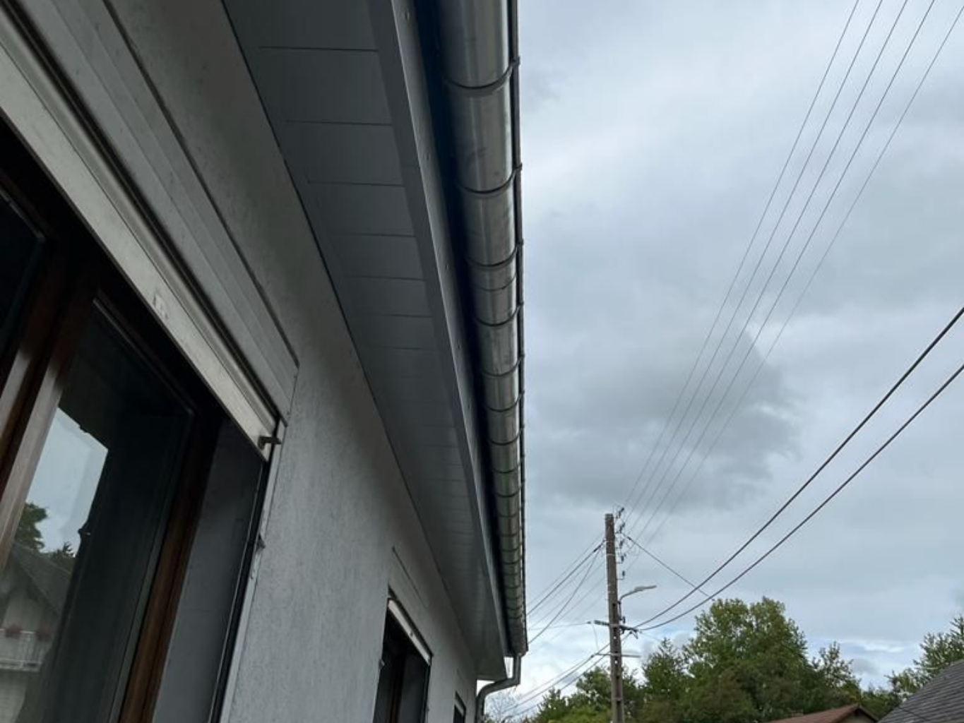 Gouttière grise sur un bâtiment blanc, se détachant sur un ciel nuageux avec des lignes électriques.