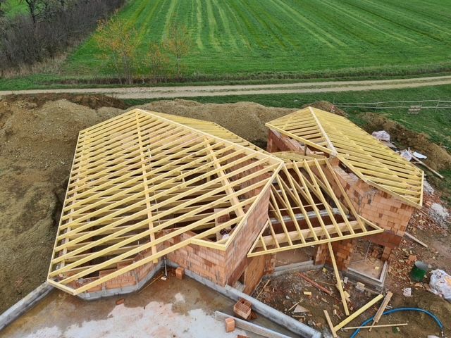 Chantier de construction avec charpente de toit en bois sur un bâtiment en briques, donnant sur un champ vert.