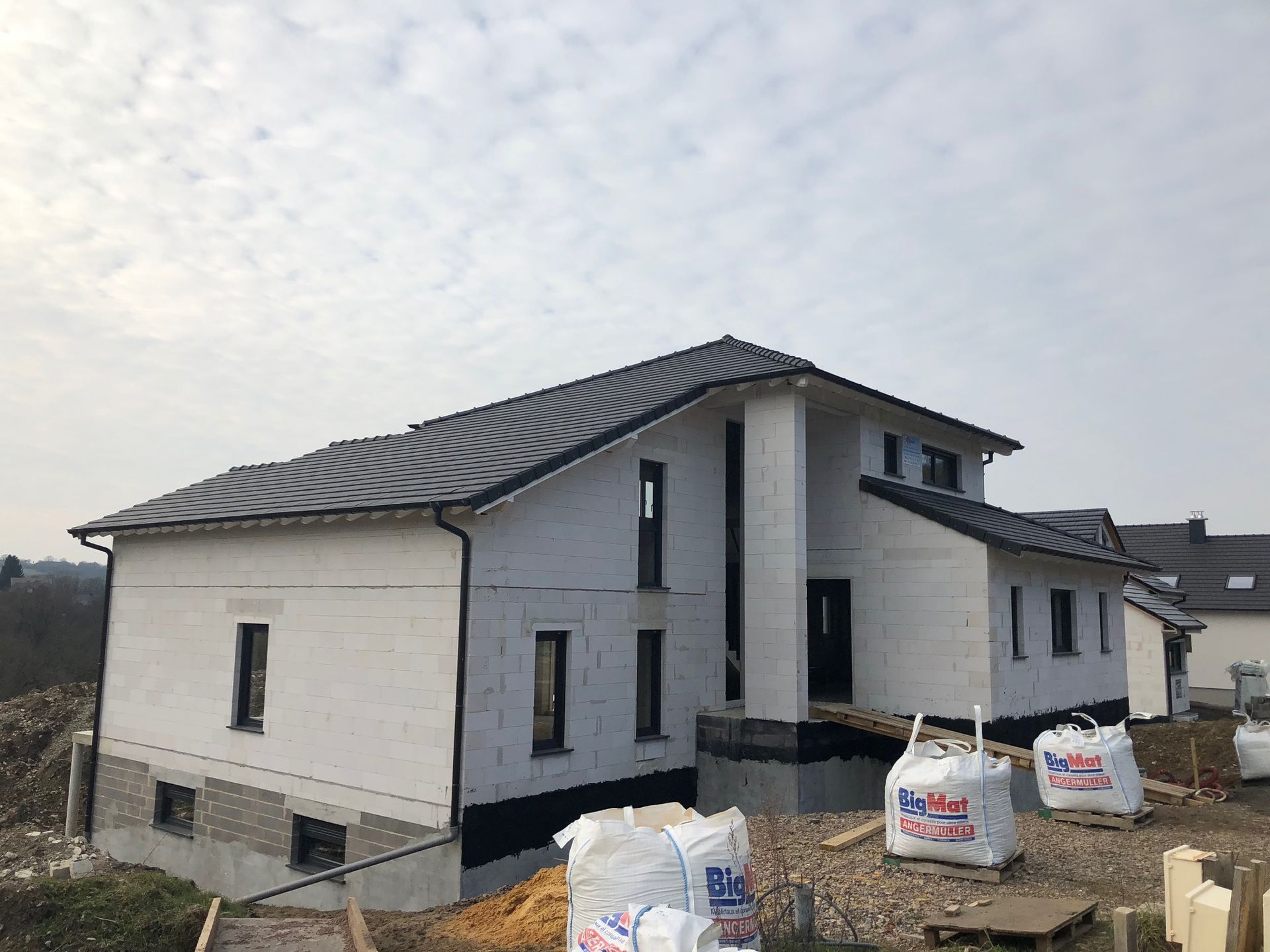 Maison en construction ; murs blancs, toit sombre, matériaux de construction visibles, ciel couvert.