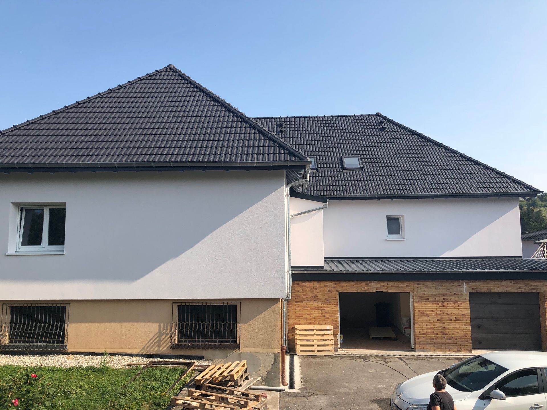 Maison à deux étages avec toit gris foncé, murs blancs et garage en briques. Une personne se tient à proximité.