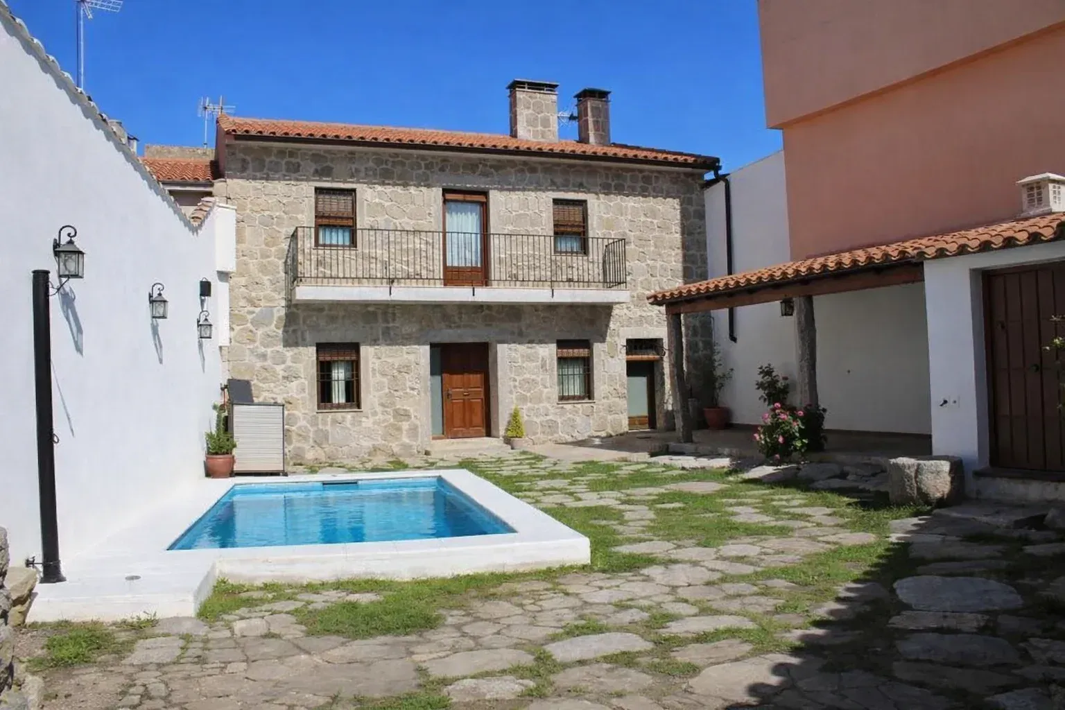 Patio de piedra con una pequeña piscina azul junto a una casa rústica de dos pisos bajo un cielo despejado.