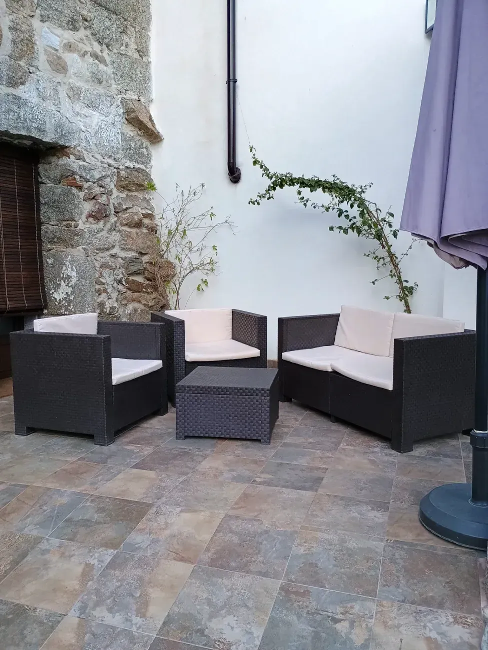 Terraza exterior con sillas de mimbre oscuro, cojines beige y una mesa pequeña contra una pared de piedra y fondo blanco.