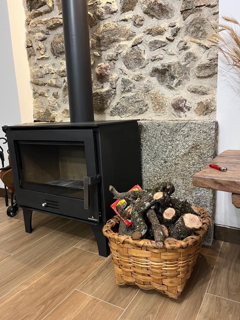 Estufa de leña negra contra una pared de piedra junto a una cesta de mimbre con leña sobre un suelo de madera.