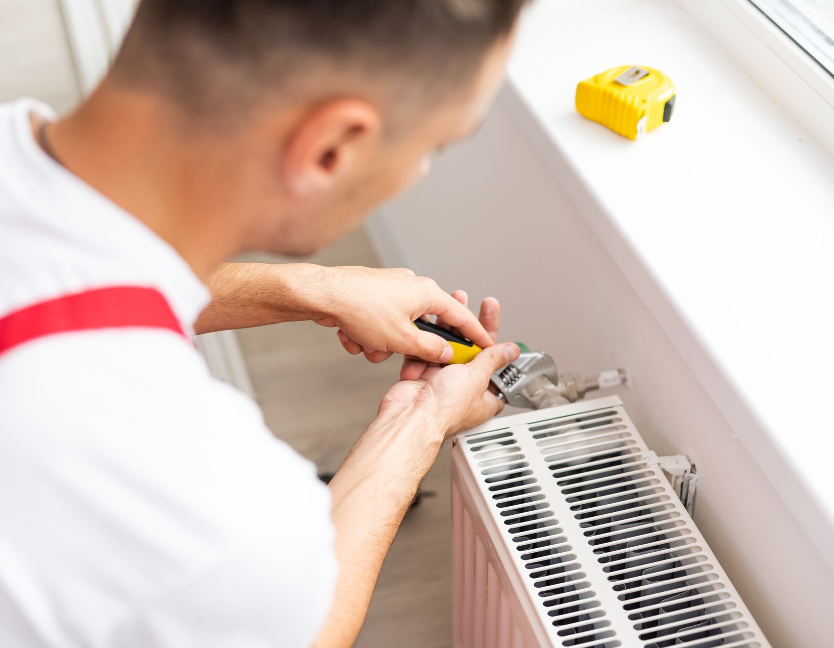 Un homme en chemise blanche répare un radiateur avec un tournevis ; un ruban à mesurer jaune sur le rebord de la fenêtre.