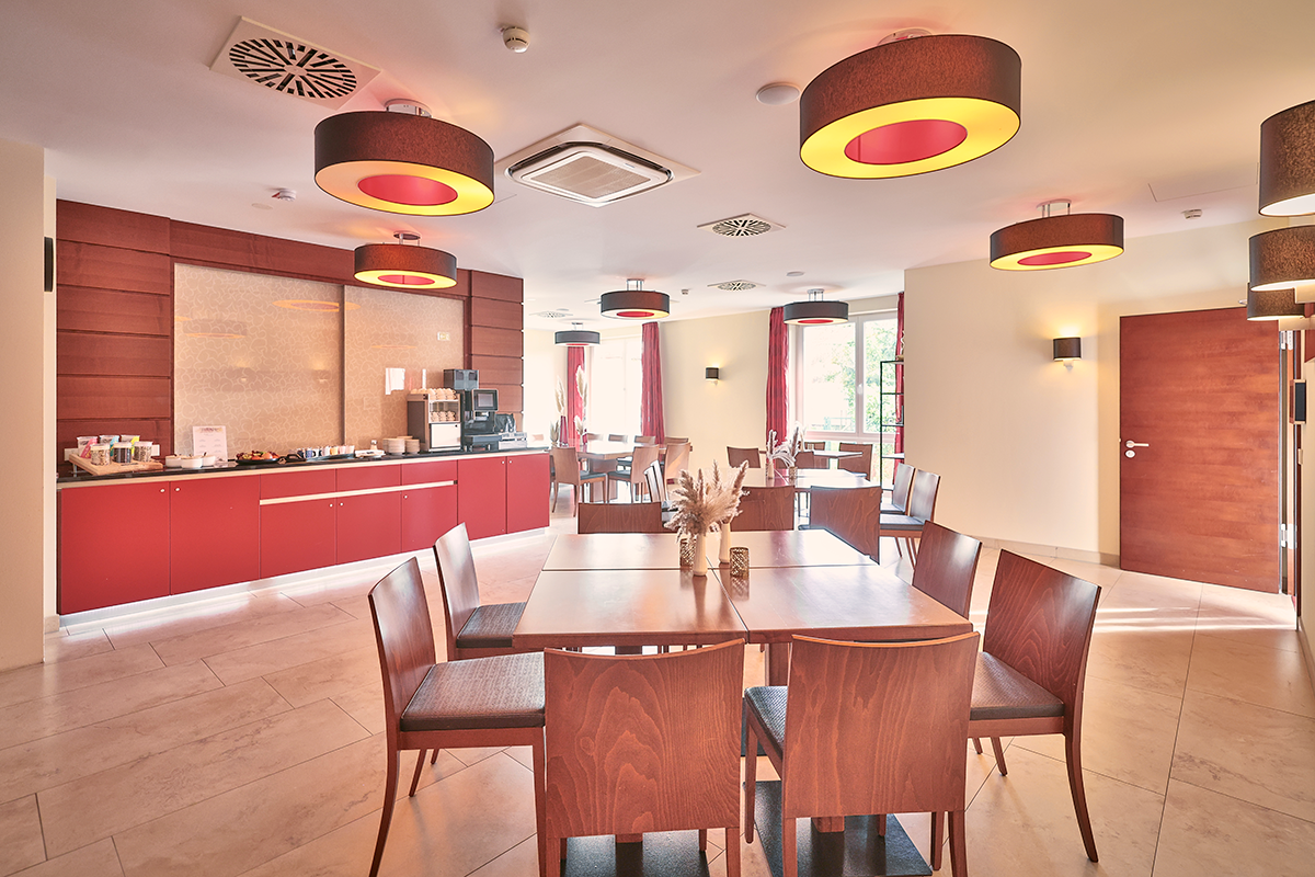 Hotelspeisesaal mit langen Holztischen, roter Buffettheke und runden Lampen.