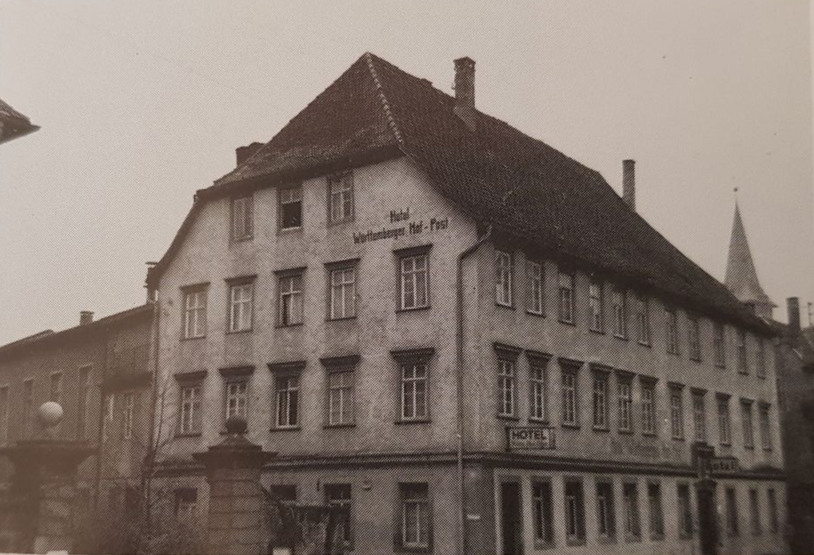 Dreistöckiges Gebäude mit schrägem Dach, Schild „Hotel“ sichtbar. Mit sichtbaren Fenstern und einem nahegelegenen Kirchturm.