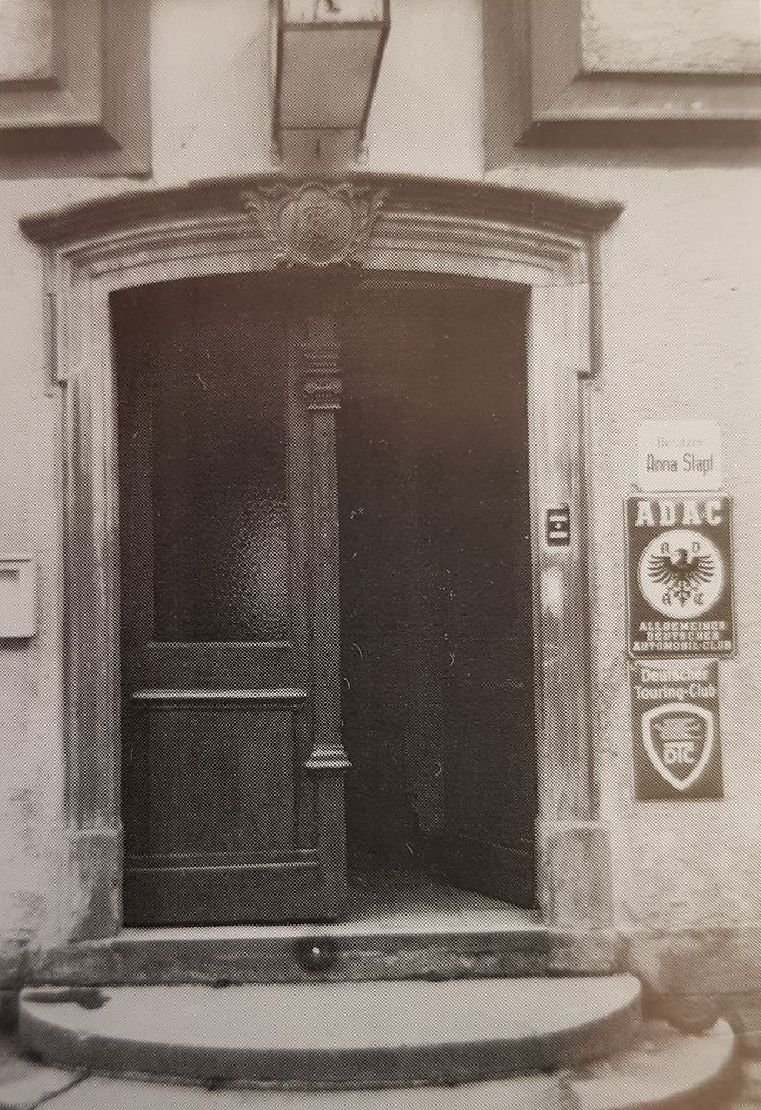 Verzierter Hauseingang mit teilweise geöffneter Holztür; Schilder für „ADAC“ und andere Dienste.