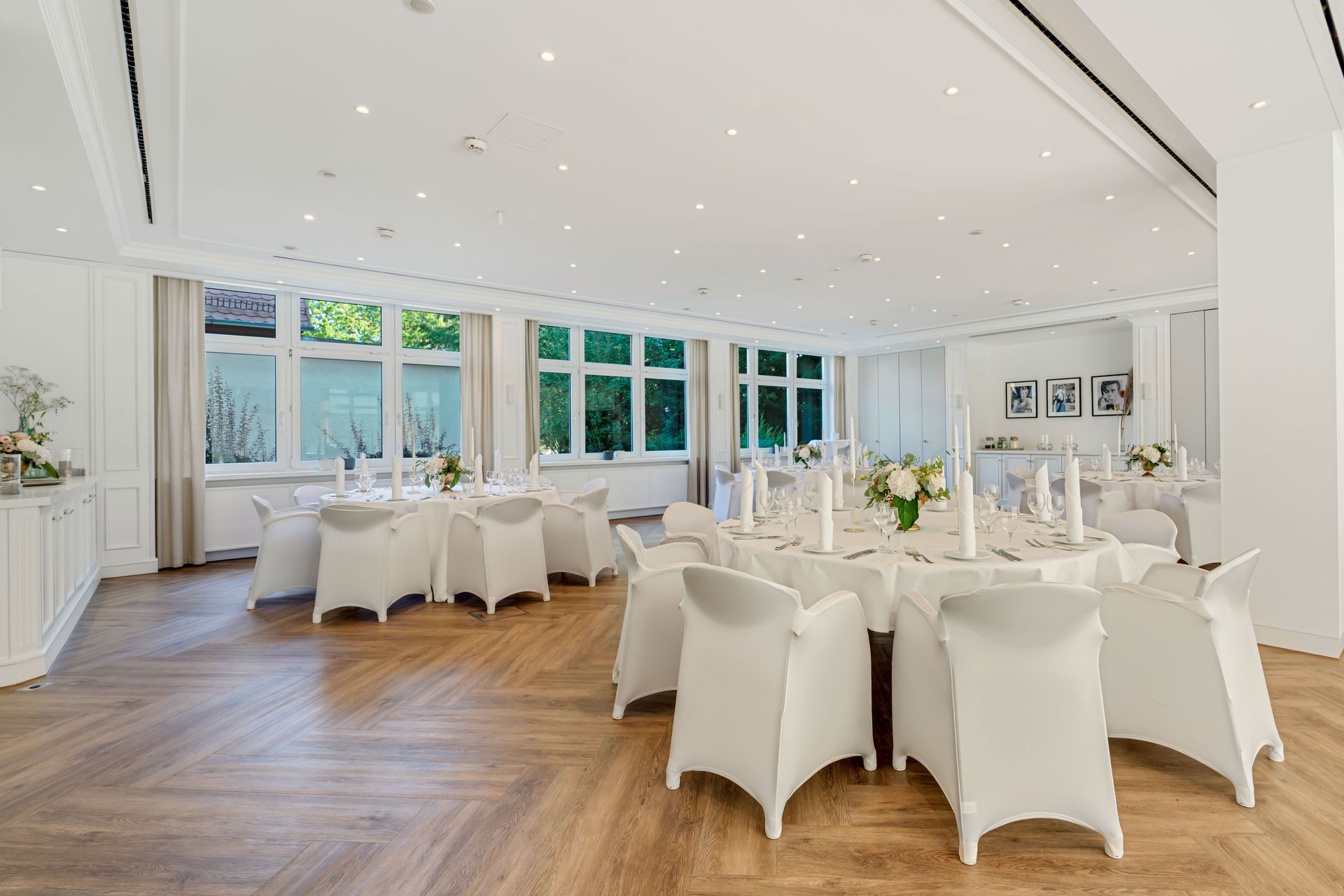 Elegantes Esszimmer mit runden Tischen für ein geselliges Beisammensein. Weiße Stühle, Blumengestecke und große Fenster.