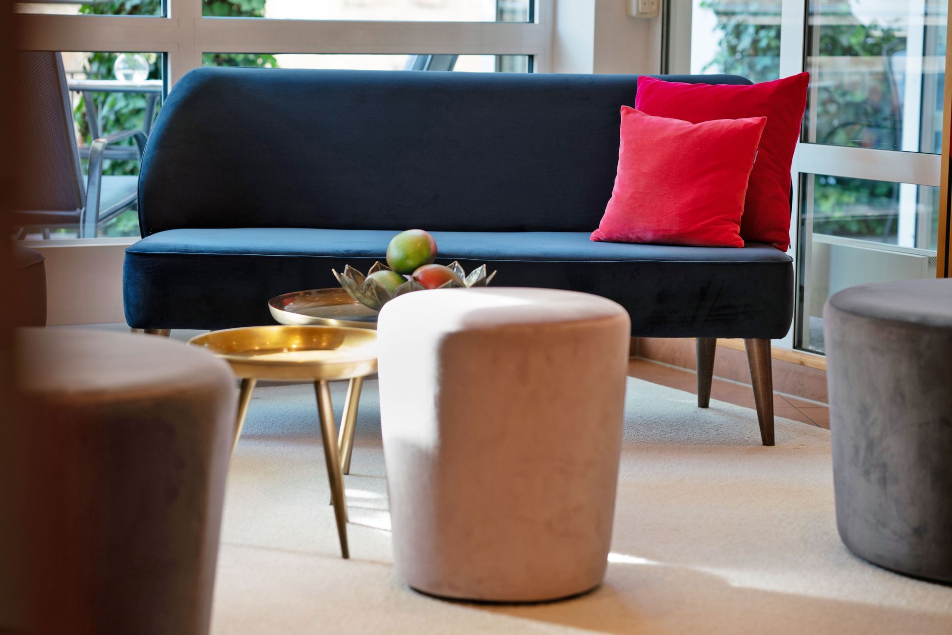 Marineblaues Sofa mit roten Kissen, kleiner goldener Tisch mit Obst und zwei runde graue Ottomanen in einem Raum mit Fenster.