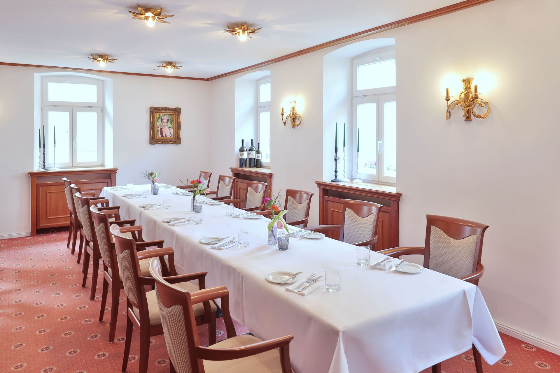 Formeller Speisesaal mit langem weißen Tisch, Stühlen, Fenstern und dekorativen Wandleuchtern.