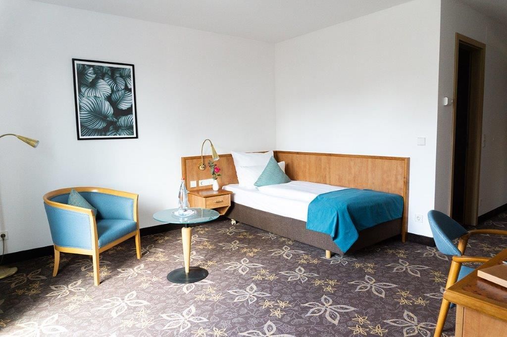 Hotelzimmer mit Einzelbett, blauen Stühlen und geblümtem Teppich.