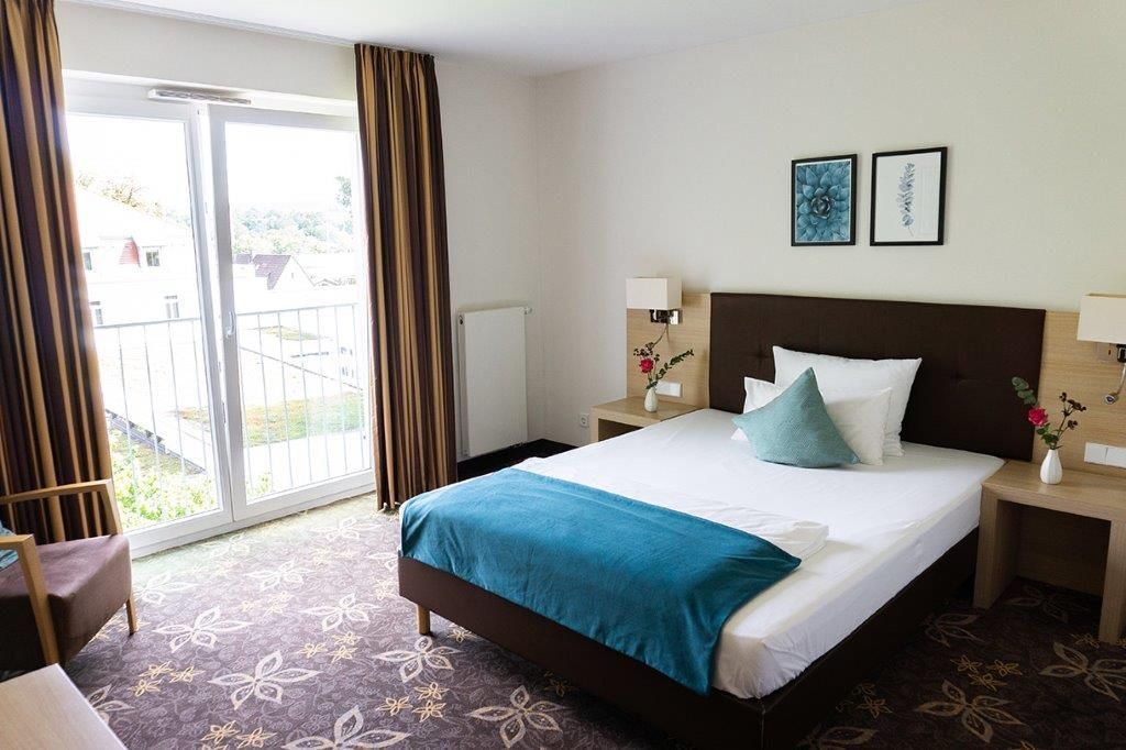 Hotelschlafzimmer mit Bett, Balkon und Stuhl; neutrale Wände, braune Vorhänge und Teppich, blaugrüne und weiße Bettwäsche.